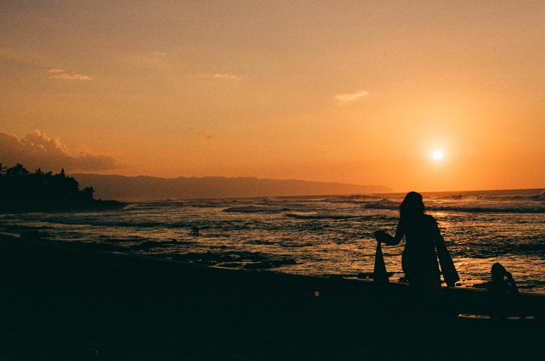 Silhouetted figures on a Hawaiian beach at sunset with waves and distant mountains
