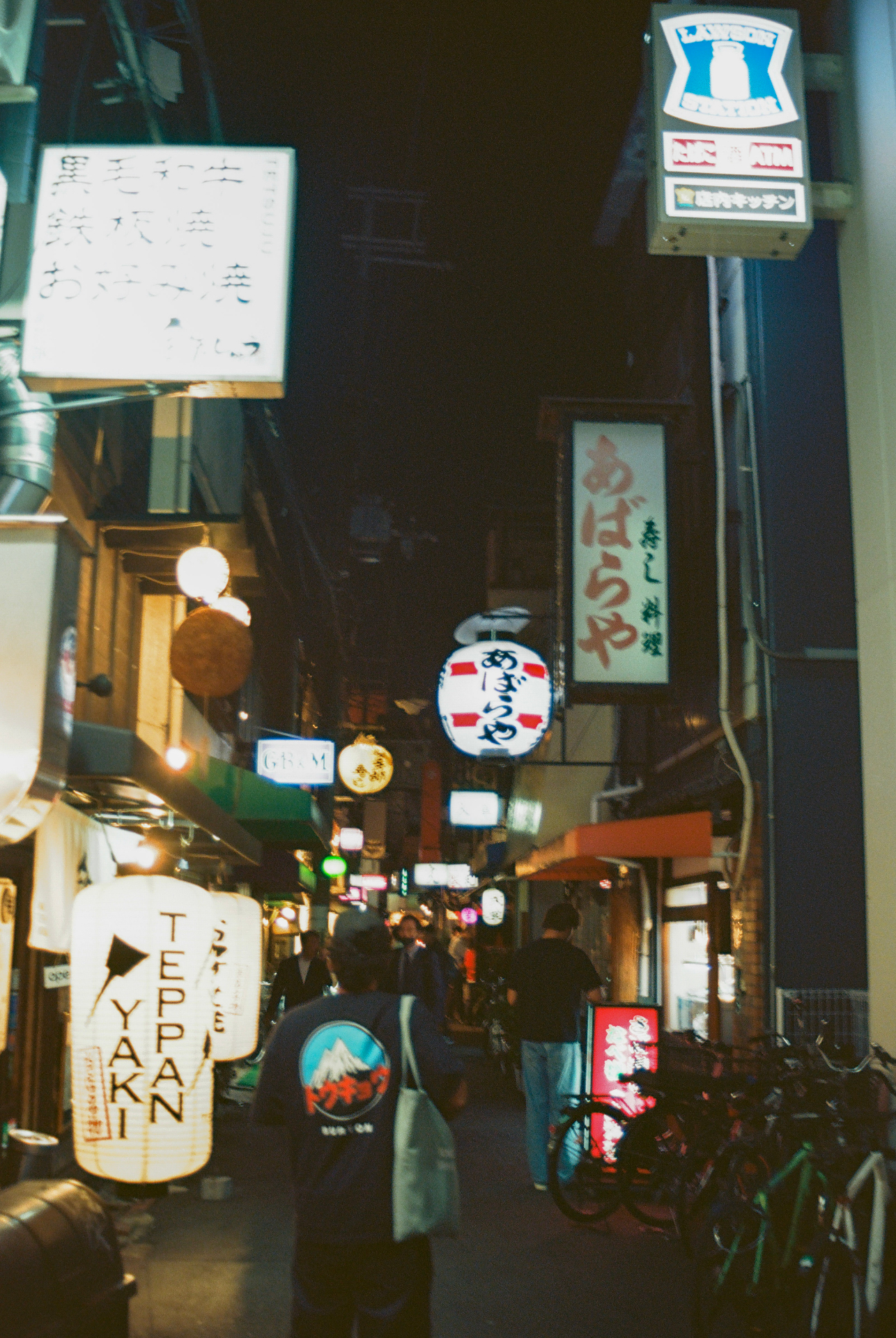 Narrow Japanese alley at night filled with paper lanterns, izakaya signs and a Lawson