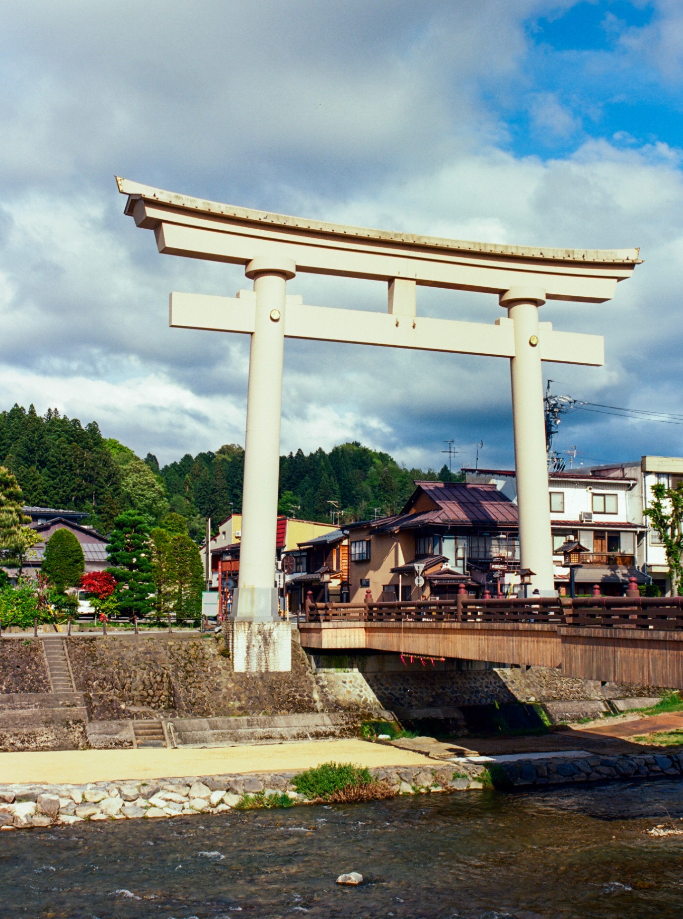 A large white torii gate above a wooden bridge over a mountain stream in Takayama