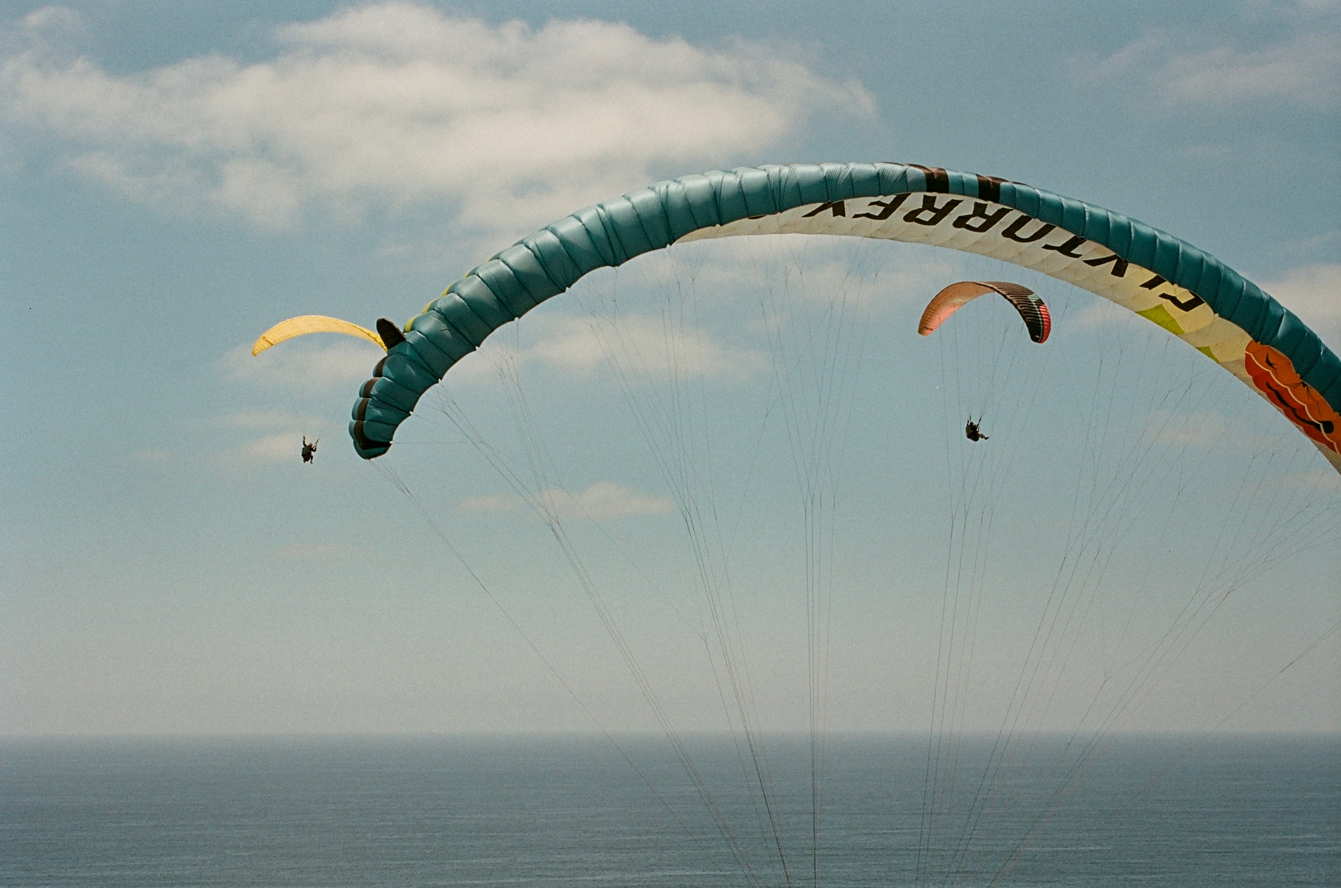 Teal Fly Torrey paraglider arcing overhead as two smaller gliders float above the ocean horizon