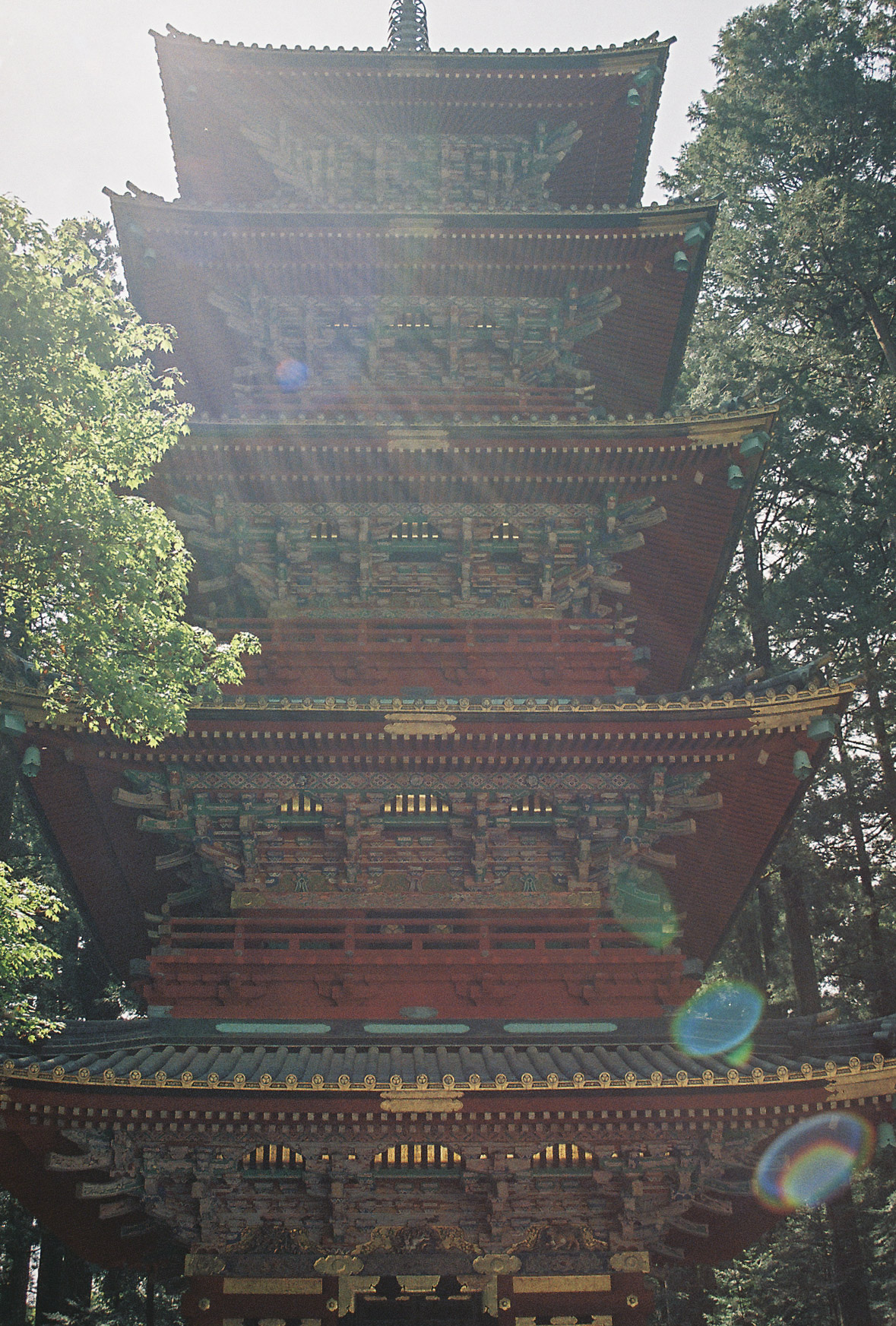 Five-story vermilion and gold pagoda rising through cedar trees in Nikko, lens flare in afternoon sun