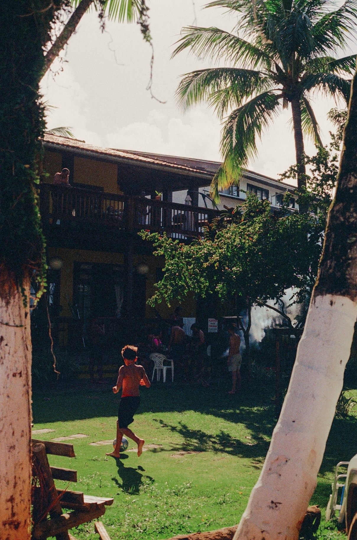 A boy running with a soccer ball on a lawn framed by palm trees and a yellow pousada