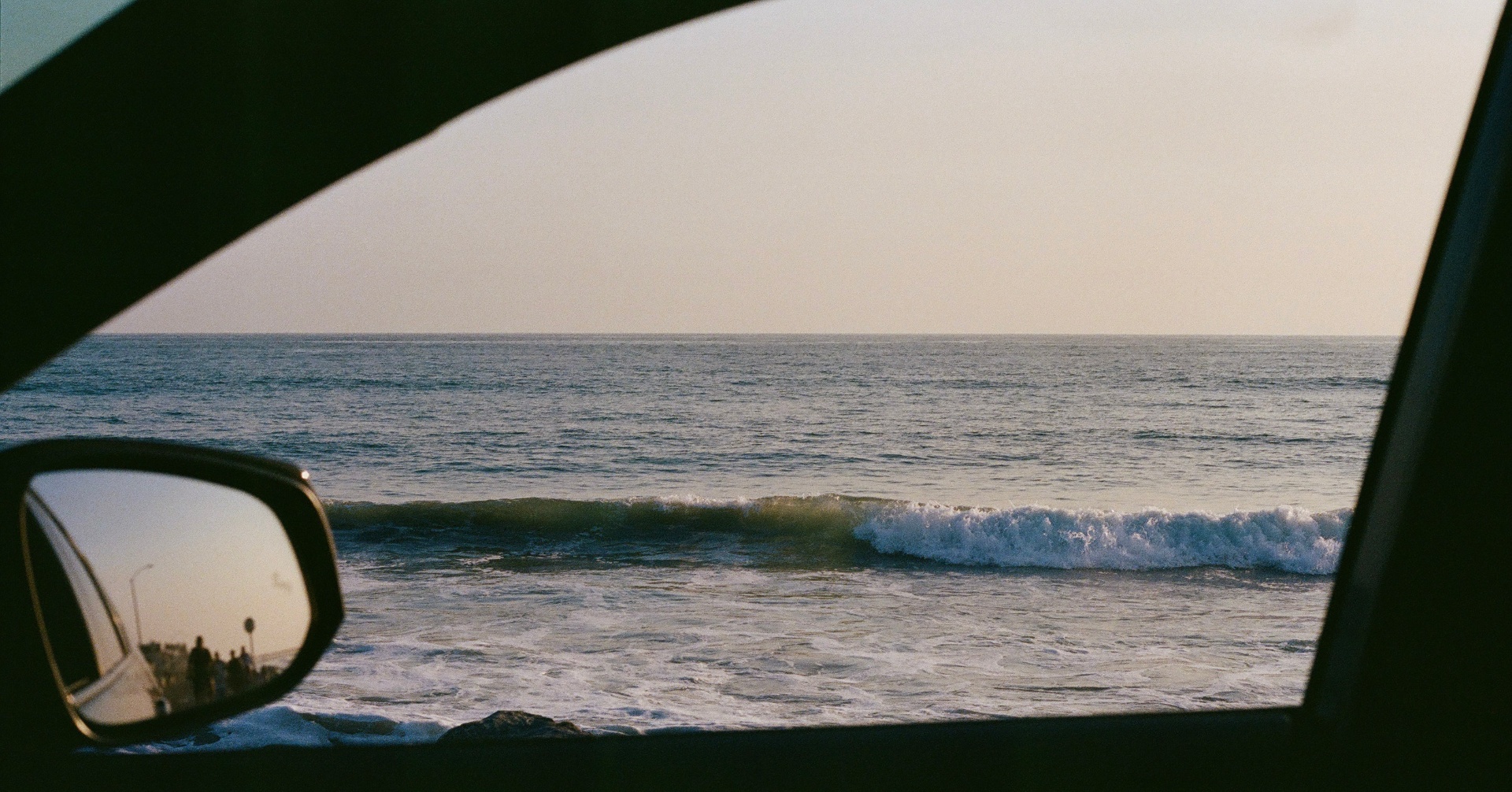 Ocean waves seen through a car window with the side mirror reflecting people by the shore