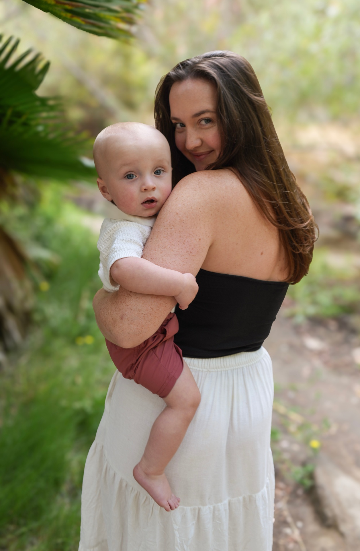 Mother holding her baby beside a palm frond in soft afternoon light