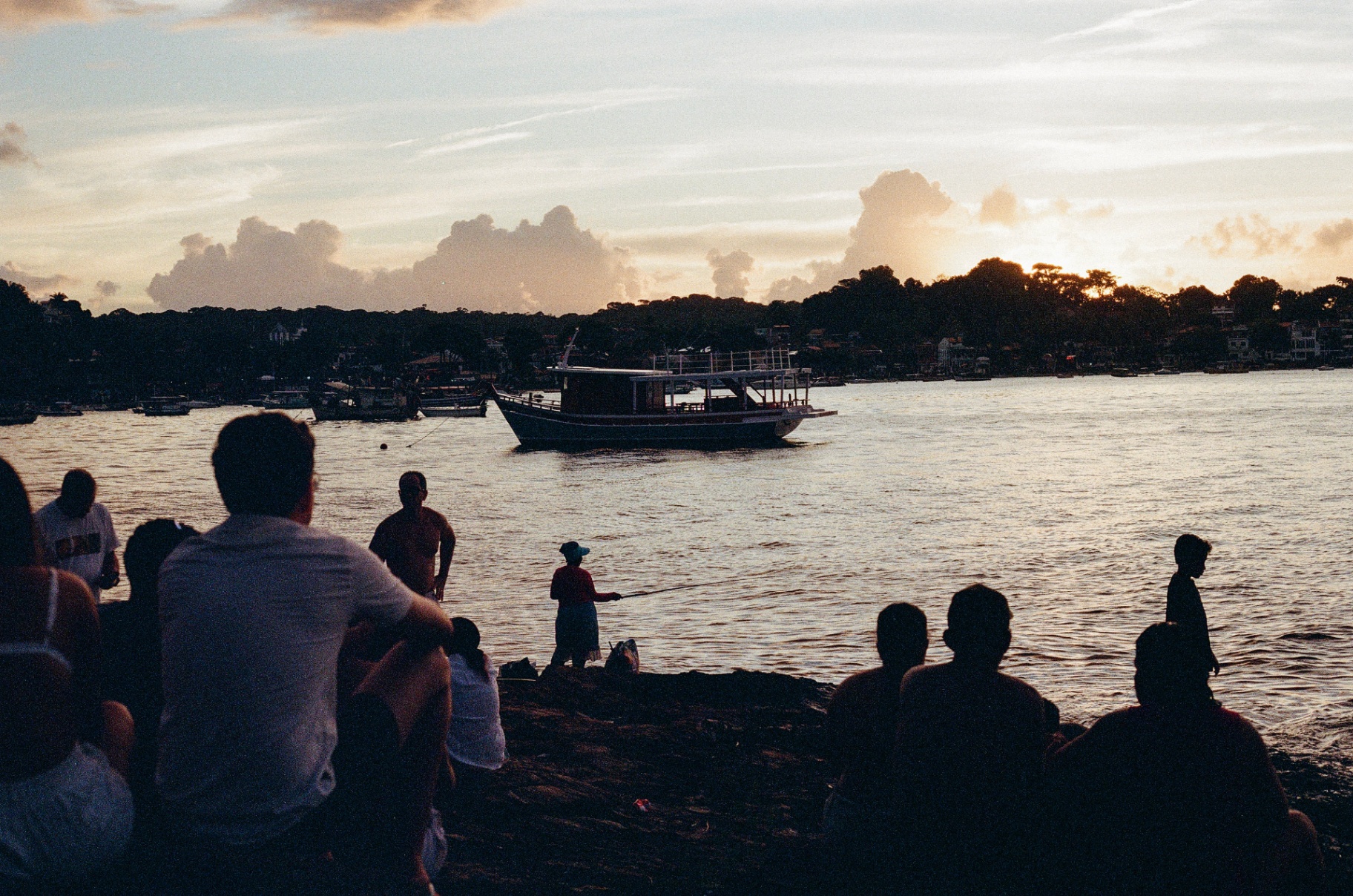 Silhouetted crowd watching a fishing boat drift across a Bahia harbor at sunset